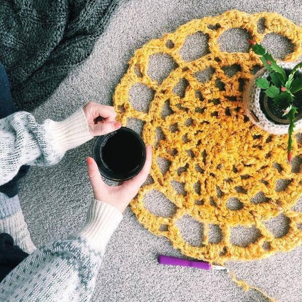 Sunshine Mat in Sunflower (yellow) on a gray surface with a houseplant, woman's hands holding coffee, and a crochet hook