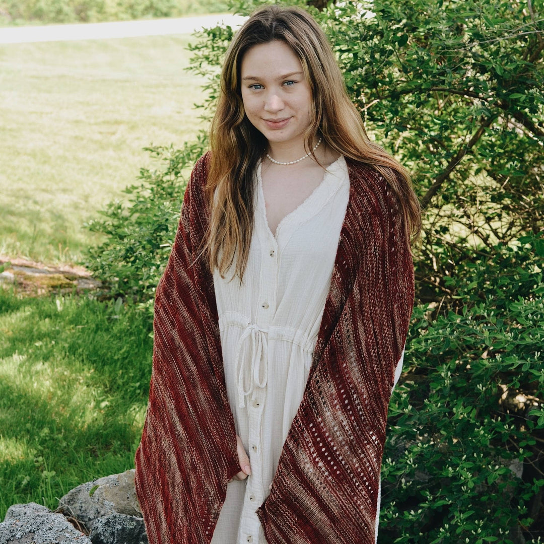 Model wearing orange and tan shawl with greenery in the background.