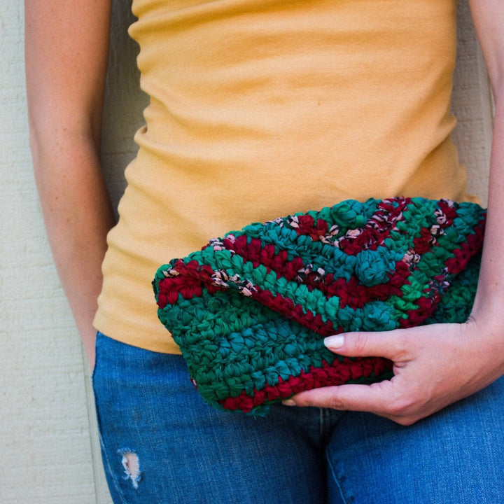 Person in yellow top holding crocheted clutch in green and red. Blue jeans visible.