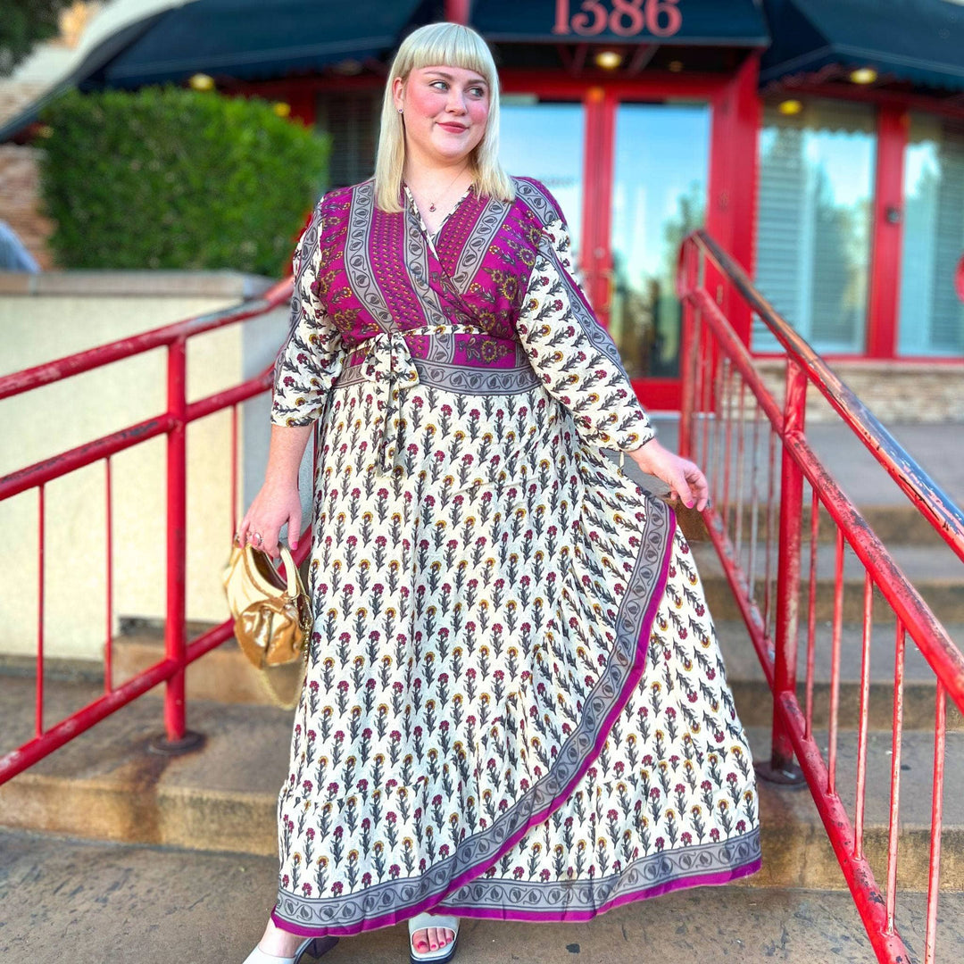 Woman on steps in a patterned maxi dress, holding a gold purse, with red doors in the background.