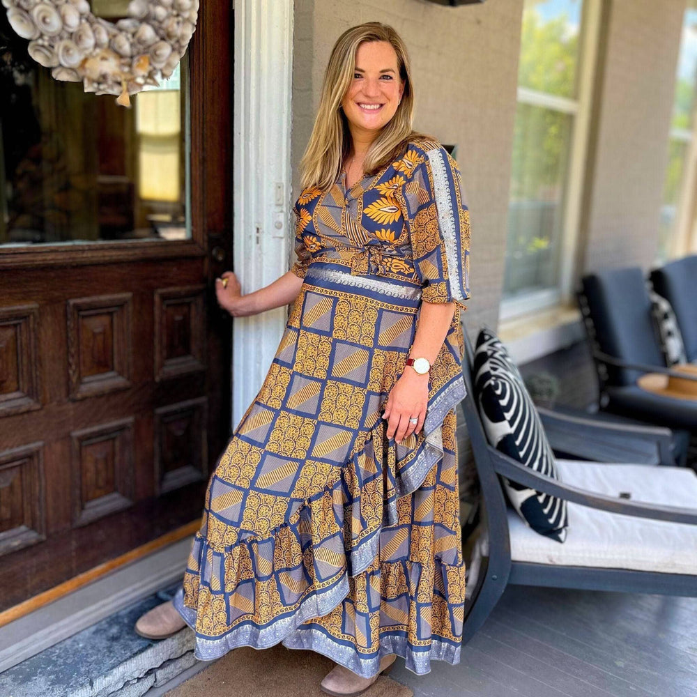 Woman in a blue and gold patterned dress, standing by a wooden door and smiling