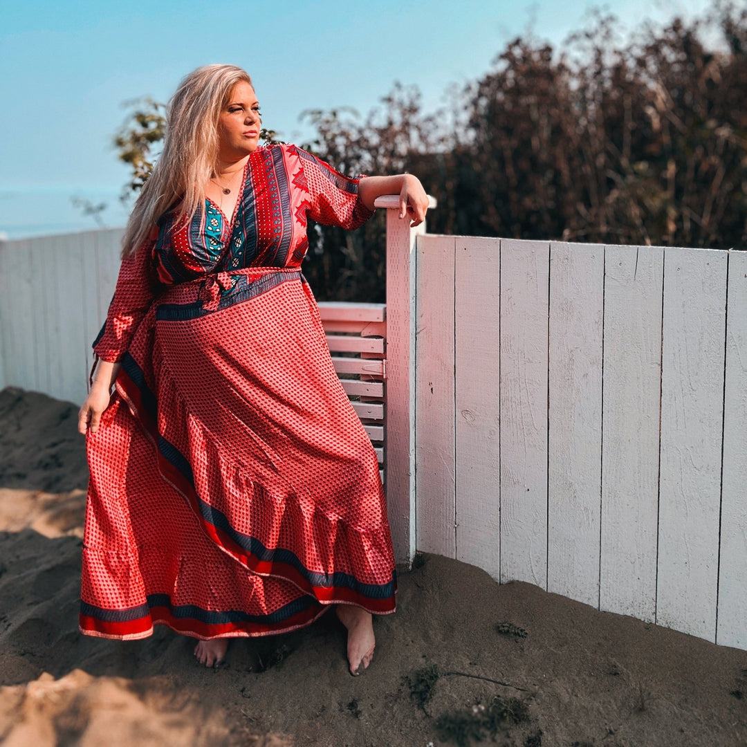 Woman in a red and black patterned maxi dress poses on a sandy beach.