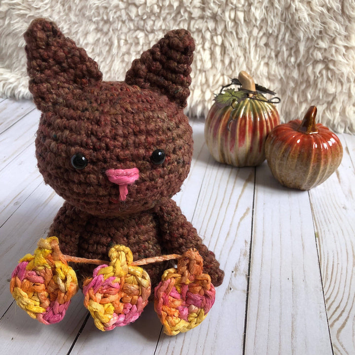 Amigurumi Crochet Cat in brown holding 3 crochet leaves sitting on a white furry surface