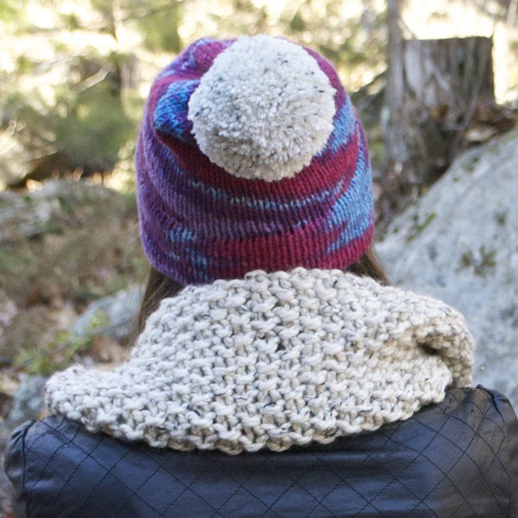 Woman wearing a purple beanie with white pom pom and white scarf and leather jacket facing a landscape background