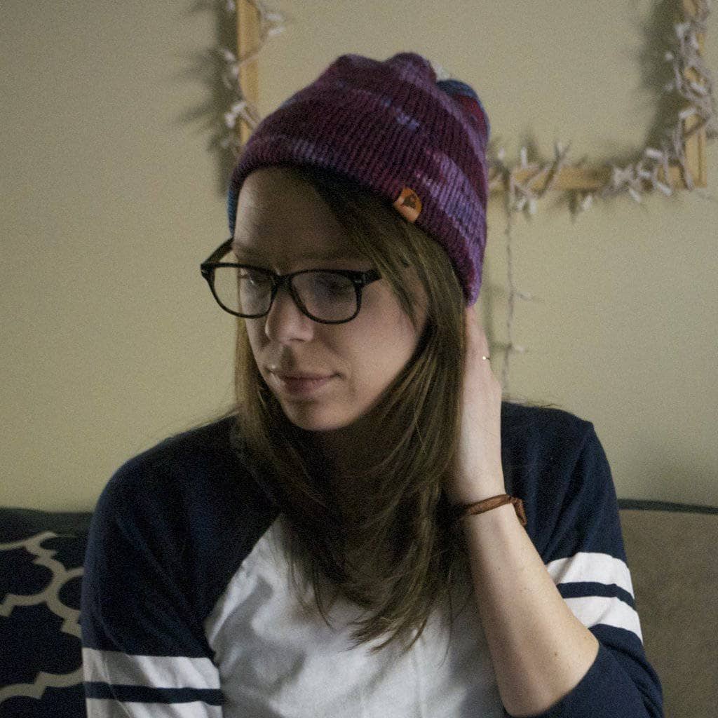 Woman wearing a purple beanie, glasses, and baseball tee sitting in front of a beige wall