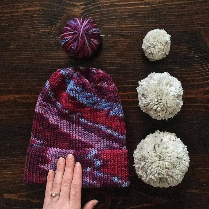 Purple beanie with white pom poms and purple yarn ball on a wooden surface