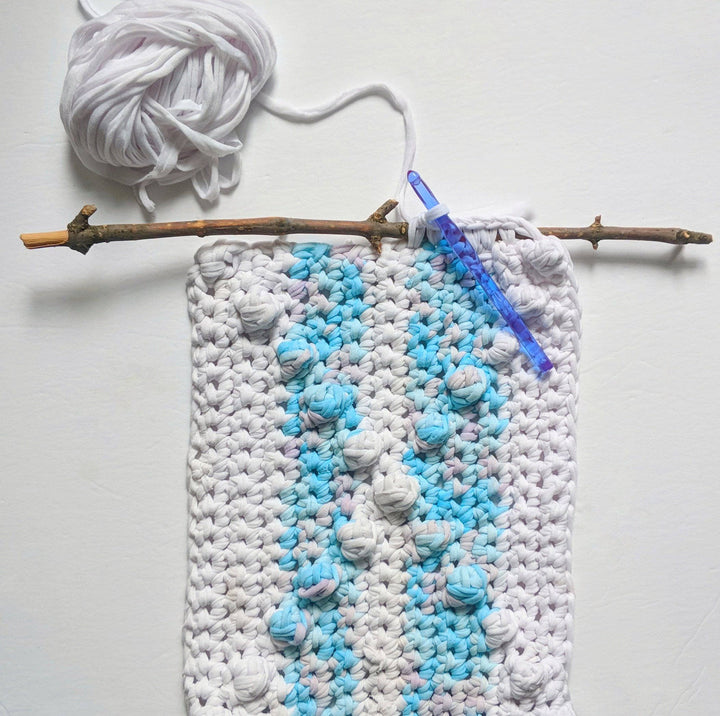 X Marks The Spot Wall Hanging in Mermaid Swirl (blue and white) hanging on a white wall next to a crochet hook and yarn ball