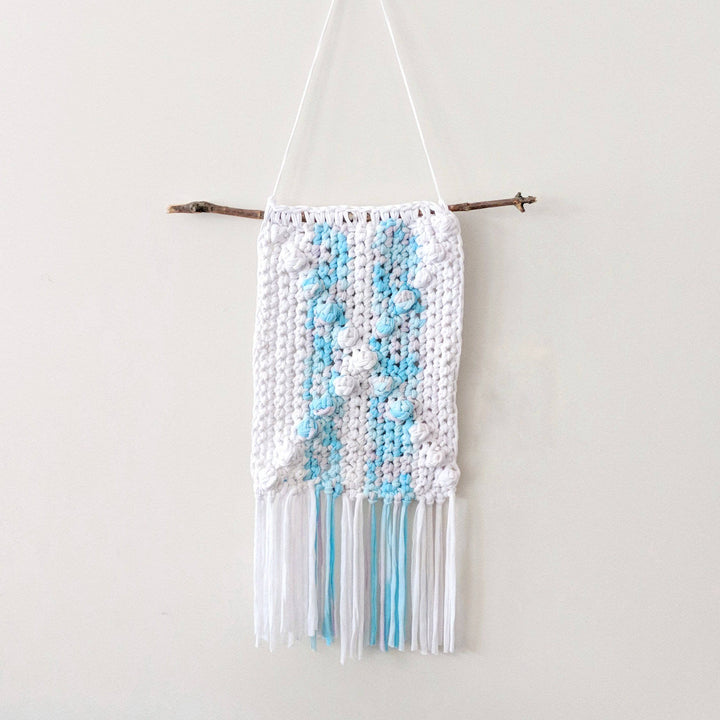 X Marks The Spot Wall Hanging in Mermaid Swirl (blue and white) hanging on a white wall next to a crochet hook and yarn ball