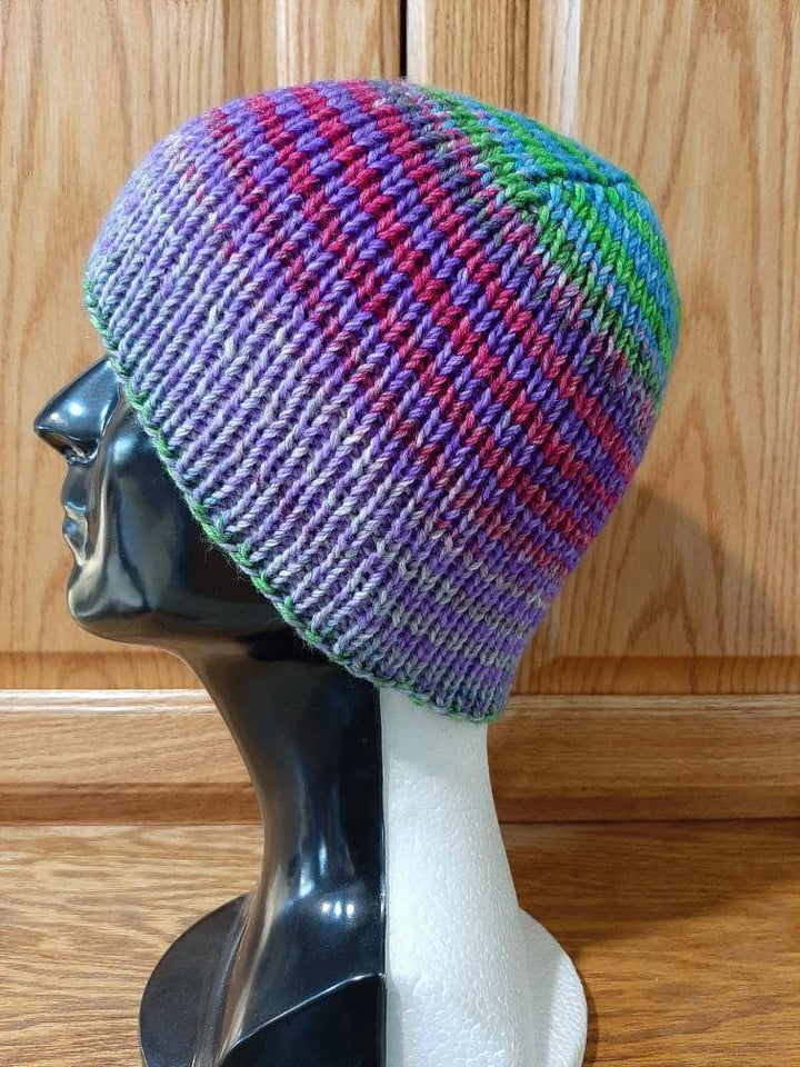 Boss beanie in alanya sock yarn, purple, white, pink, green and blue atop a black and white mannequin head in front of wood background.