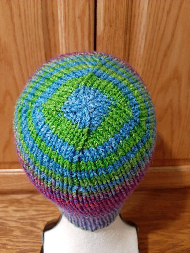 Top view of boss beanie sitting atop a mannequin head in front of a wood background. Crown is blue and green with simple spiral decreases.