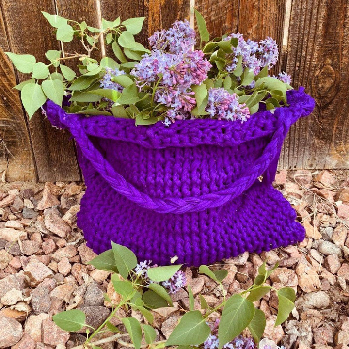 Knitted T-Shirt Bag in Pantone Ultra Violet standing in front of a wooden fence and holding purple flowers