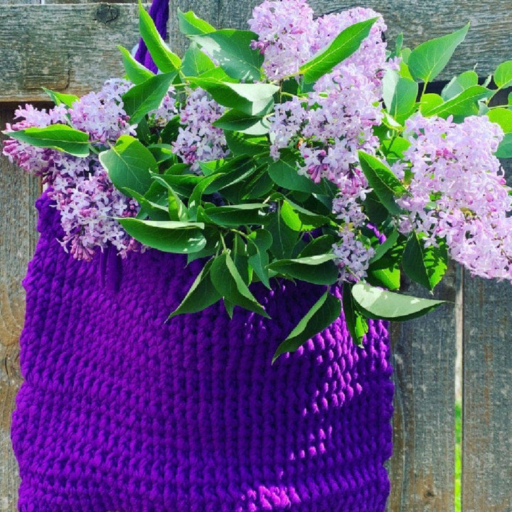 Knitted T-Shirt Bag in Pantone Ultra Violet hanging from a wooden fence and holding purple flowers