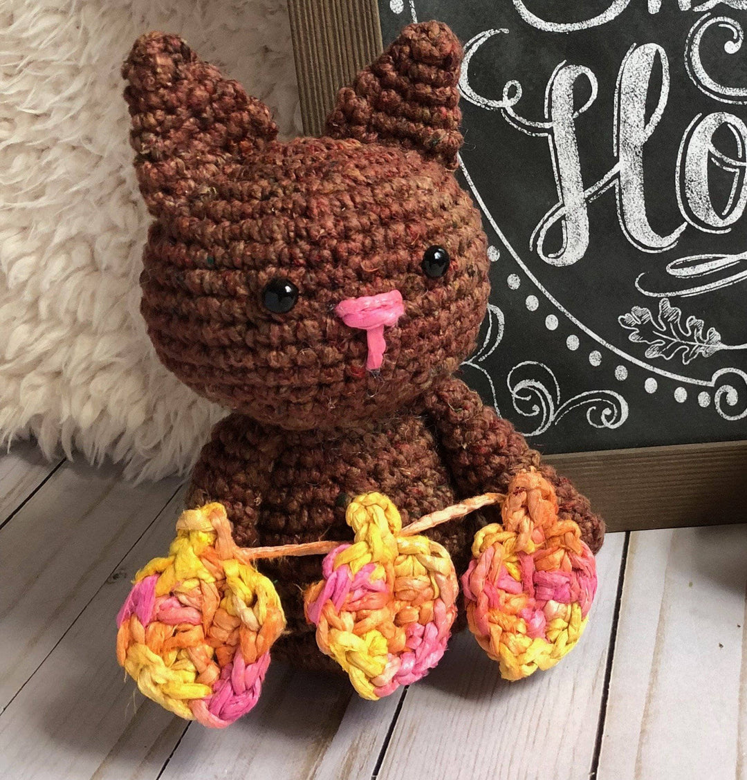 Amigurumi Crochet Cat in brown holding 3 crochet leaves in front of a chalkboard sign on a wooden floor