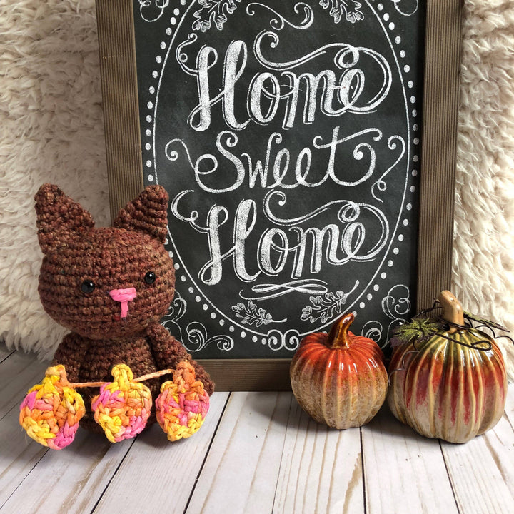 Amigurumi Crochet Cat in brown holding 3 crochet leaves in front of a chalkboard sign on a wooden floor